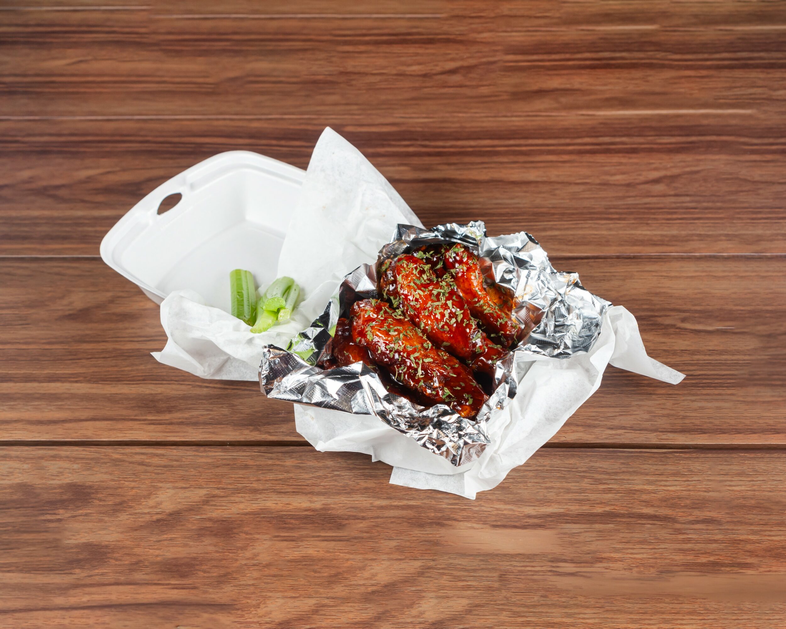 Barbecue chicken wings in foil with celery sticks in a takeout container on a wooden surface.