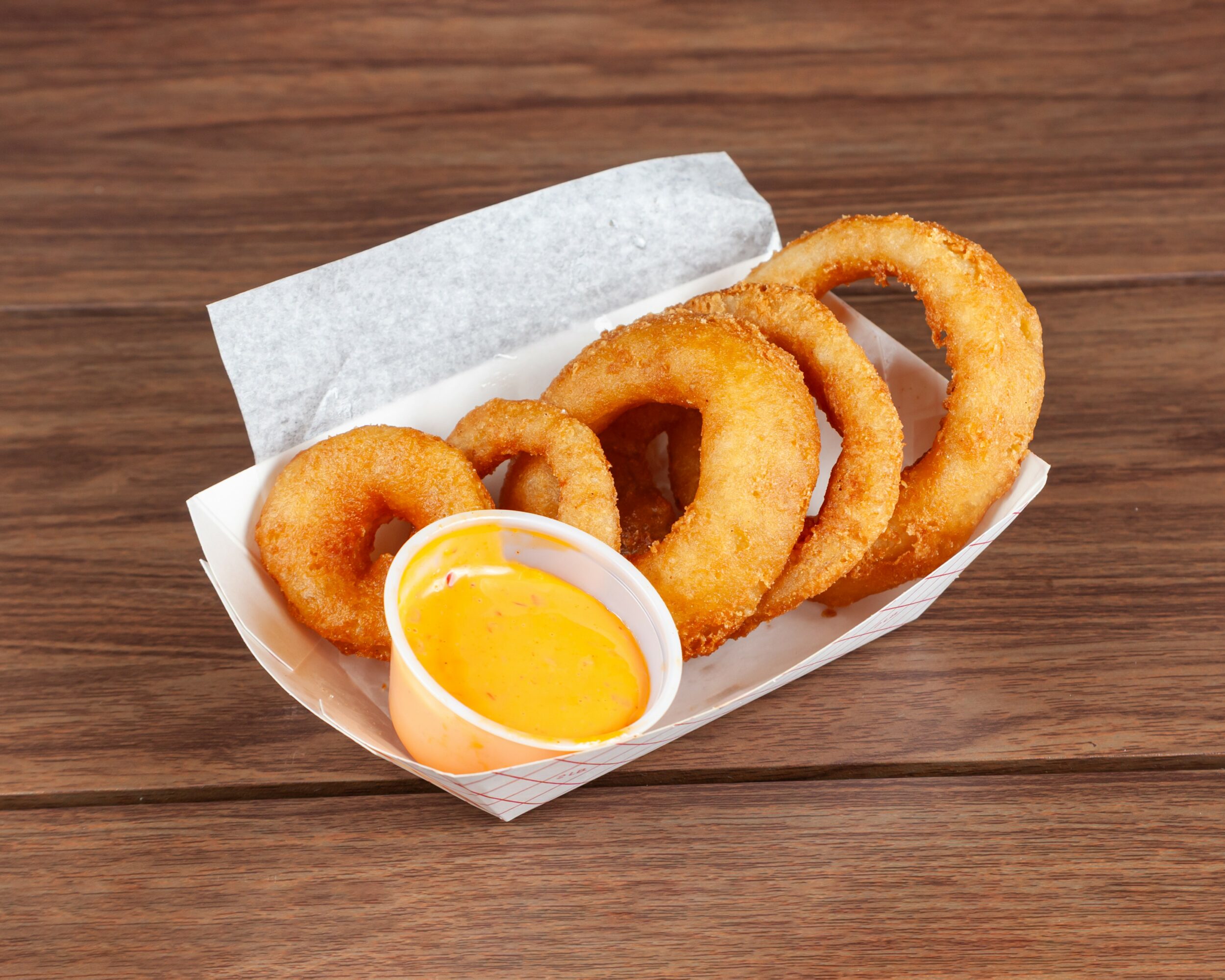 Basket with fried onion rings and a small cup of yellow dipping sauce on a wooden surface.
