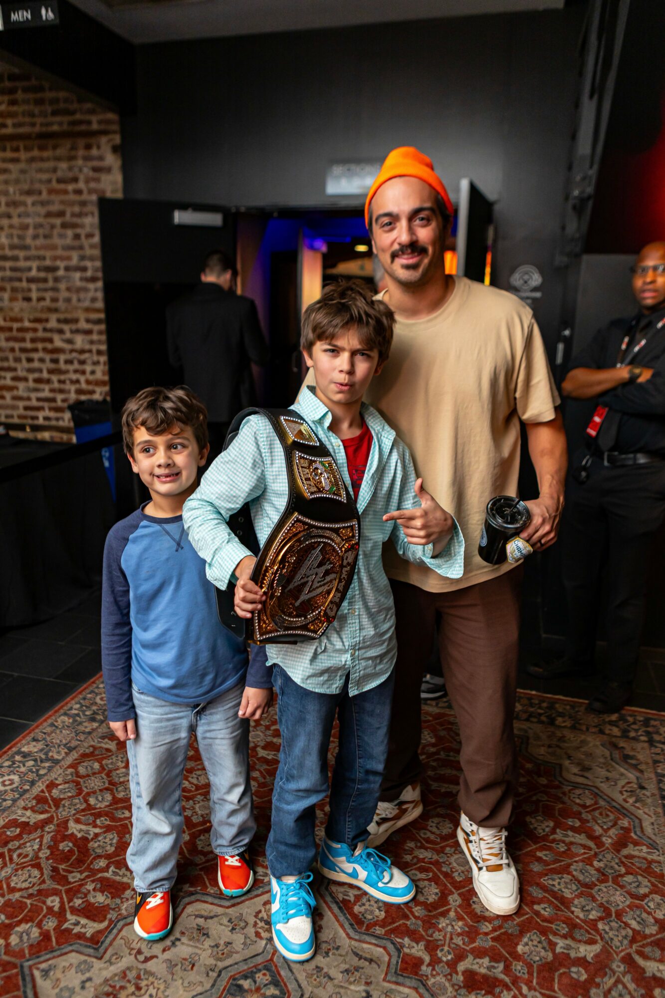 Three children and an adult man standing indoors, with a dark wall and a person in the background, smiling at the camera.