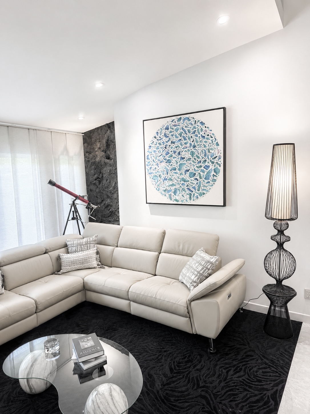 Living room with white sectional sofa, round glass coffee table, artwork, telescope, and tall lamp, with white walls and curtains.