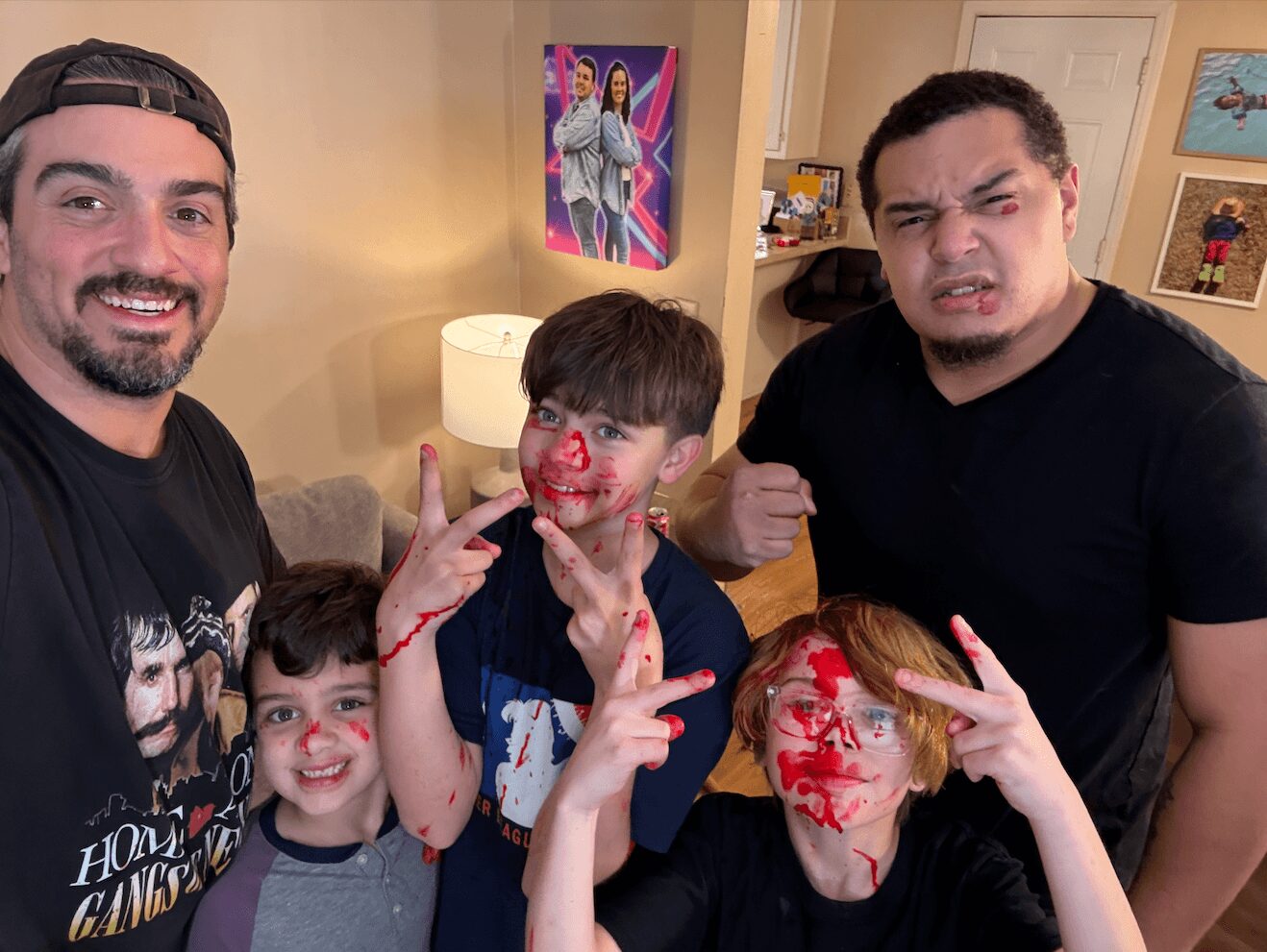 Group of five people, two adults and three children, smiling and making peace signs indoors, with colorful artwork on the wall.