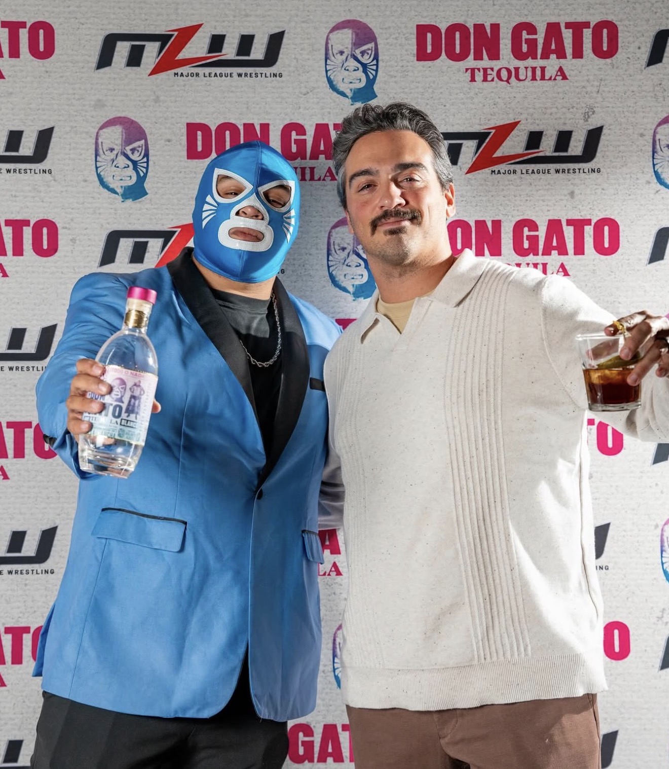 Two men posing with drinks in front of a promotional backdrop, one wearing a blue blazer and a blue luchador mask, the other in a white sweater.