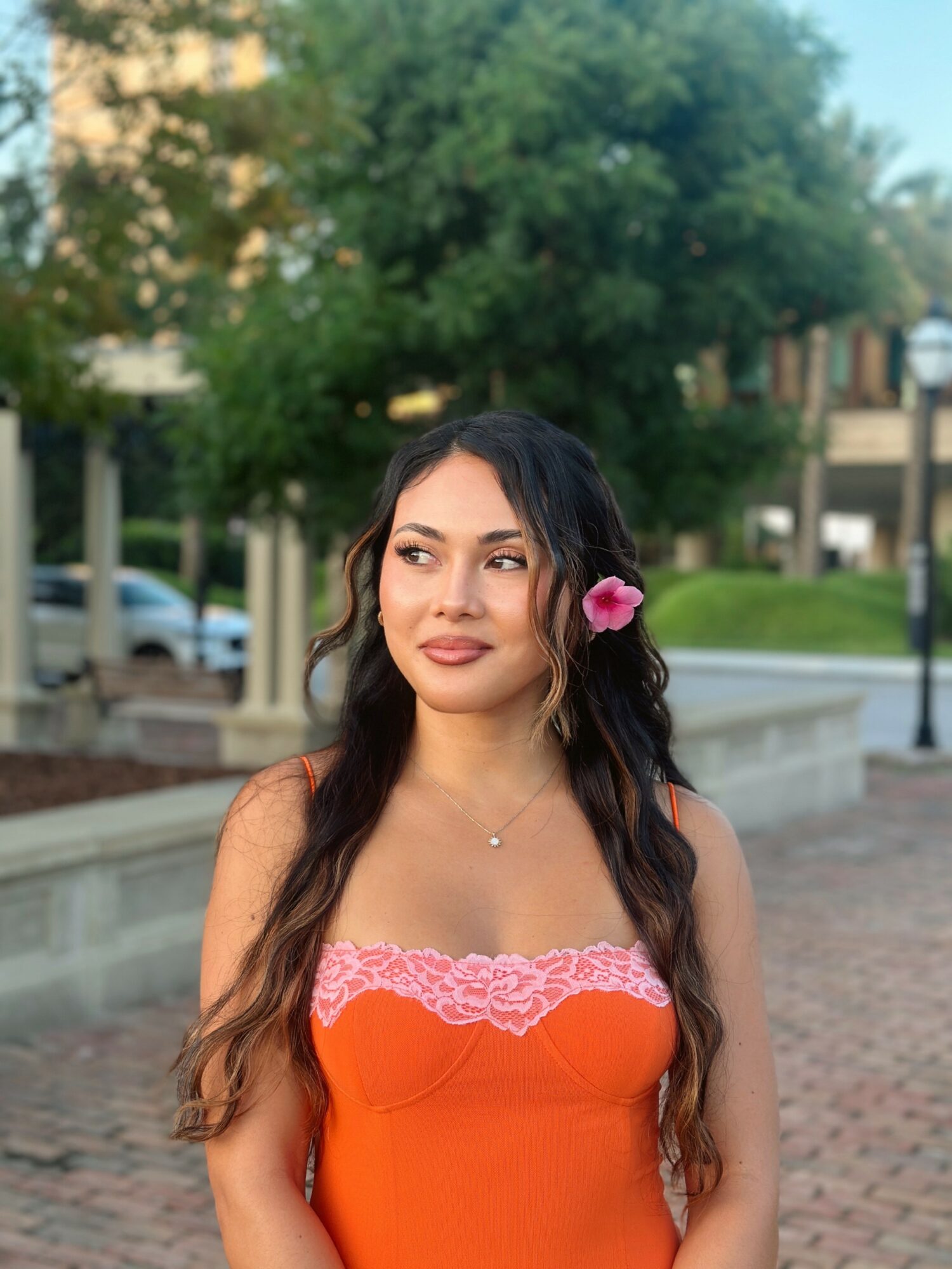 Young woman with long dark hair and a pink flower in her hair, wearing an orange strapless top, outdoors.