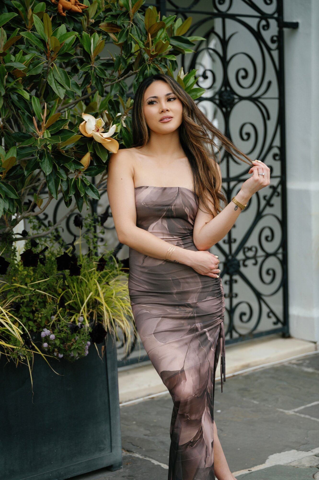 Young woman in strapless dress standing outdoors near a decorative gate and plants.