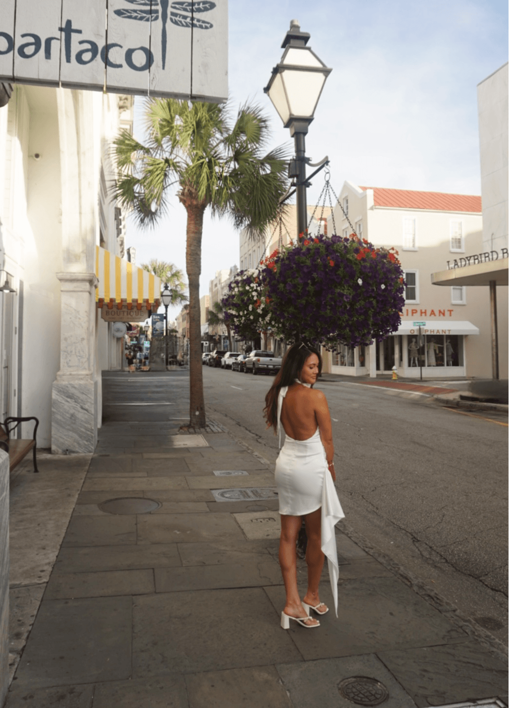 Woman in white dress and heels walking on sidewalk with palm trees and hanging flower basket nearby.
