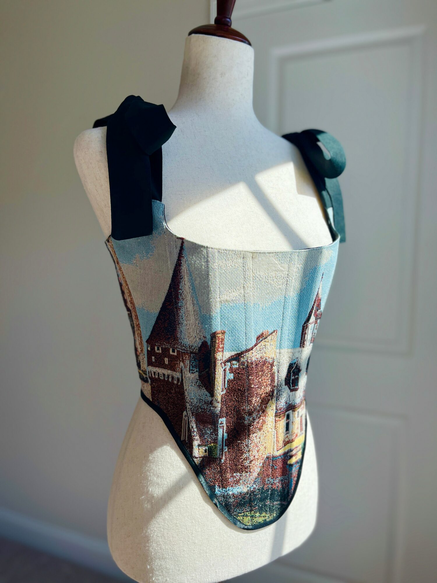 Dress with a scenic landscape print on a mannequin, sunlight casting shadows, black shoulder straps, in a room with a white door.