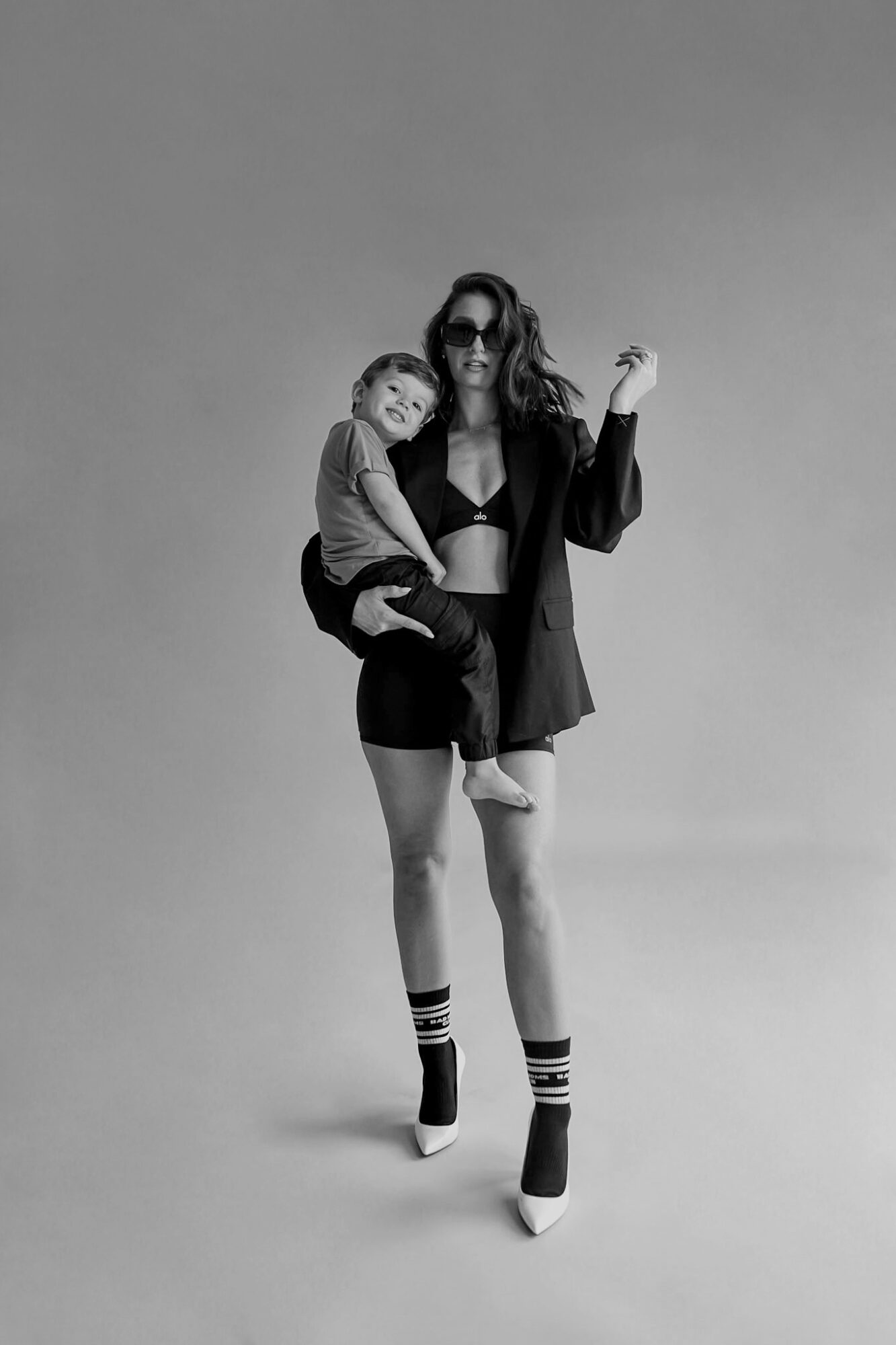 Woman wearing sunglasses and a black outfit holds a child, both smiling, against a plain background.