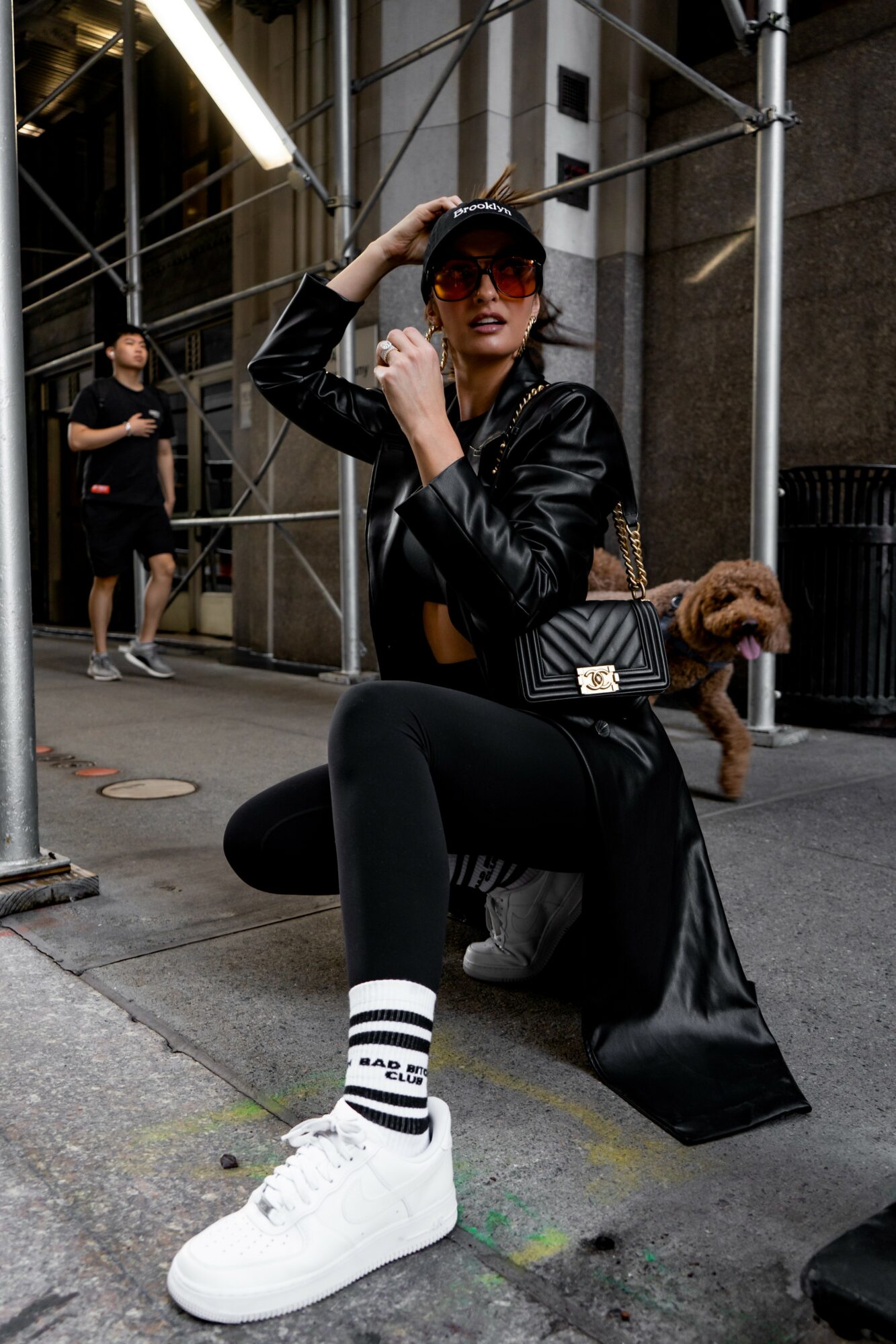 Woman sitting on the sidewalk with scaffolding behind her, wearing sunglasses, a black jacket, and white sneakers.
