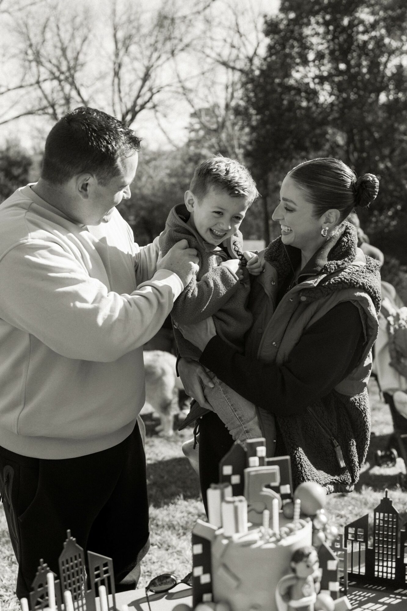 Family outdoors with a child, smiling and playing, trees in background, black and white photo.