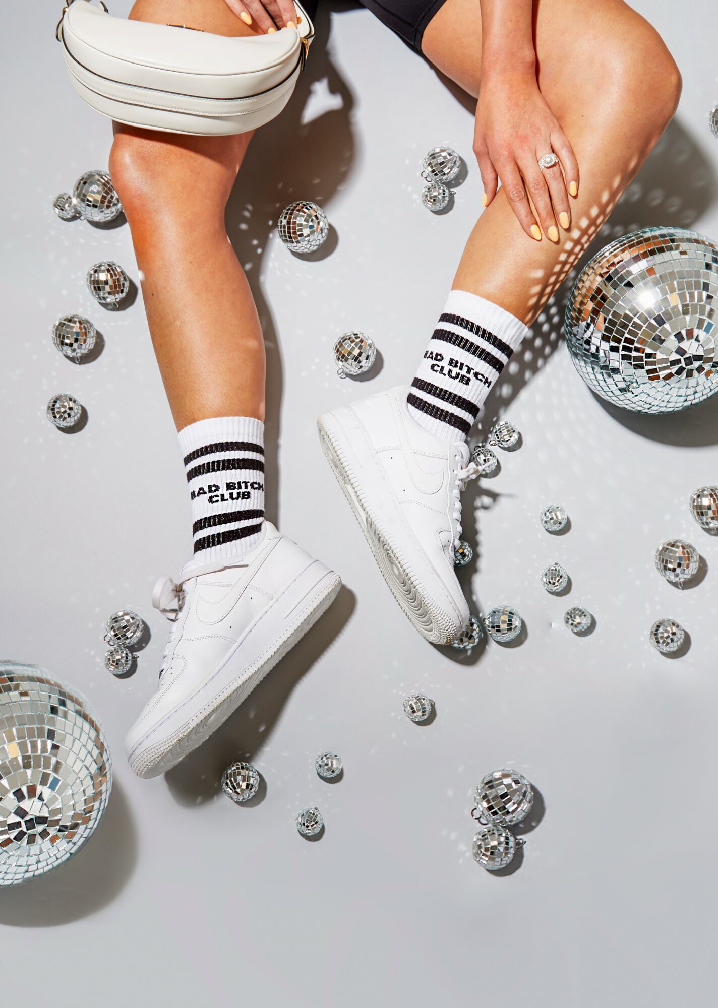 Two people wearing white sneakers and striped socks, surrounded by disco balls and small reflective spheres, on a light gray background.