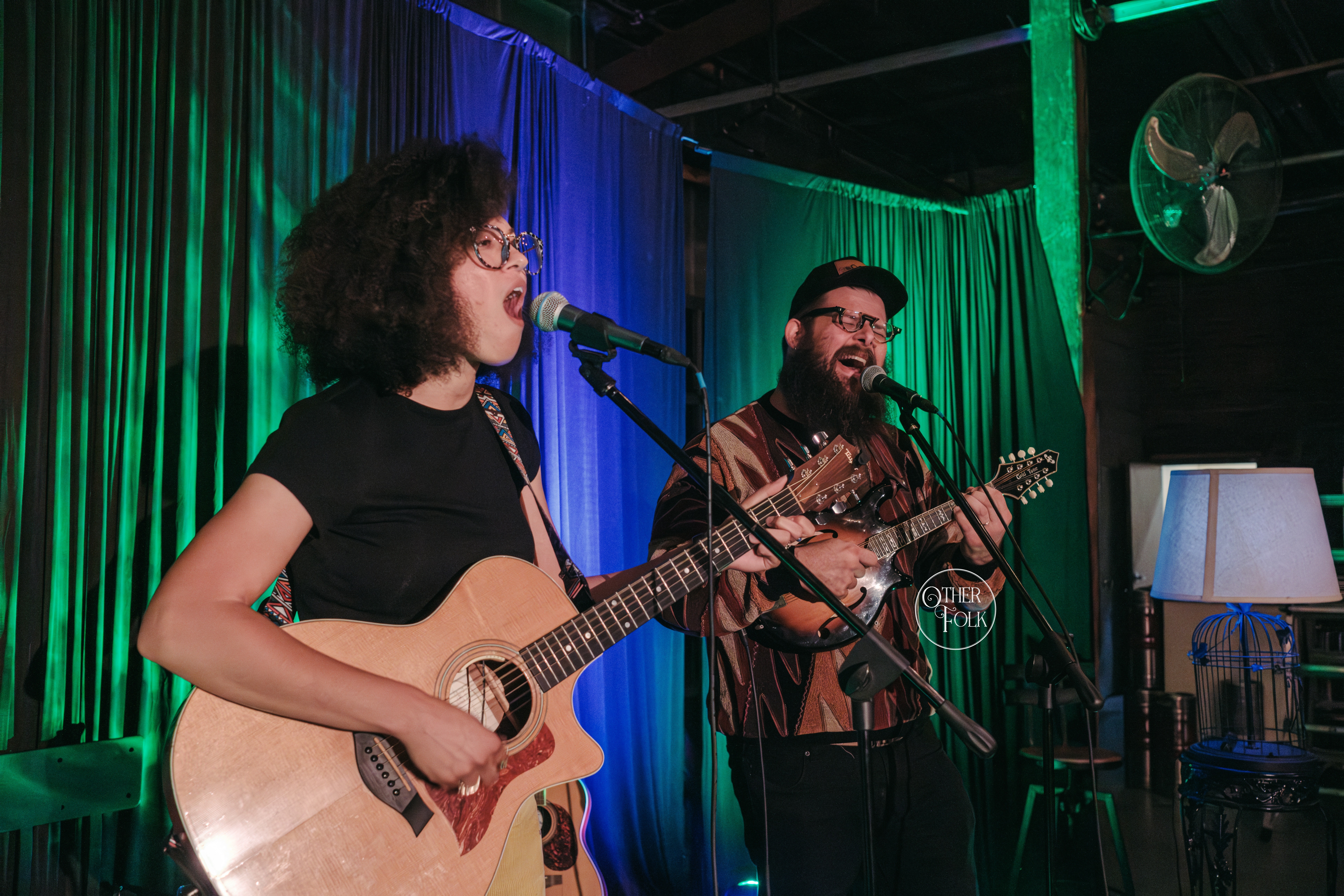 Two musicians perform on stage with guitars, colorful curtains, and a fan in the background.