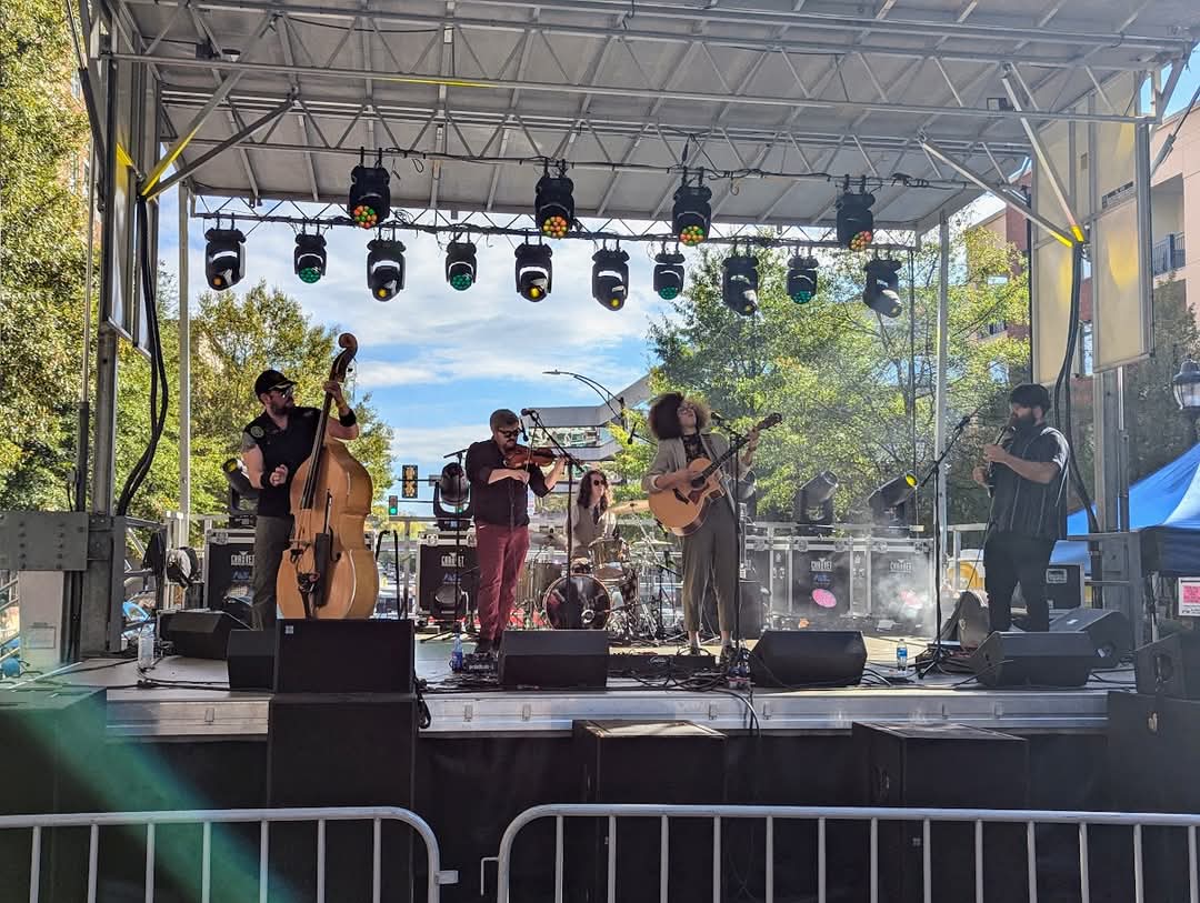Band performing on an outdoor stage with four musicians, trees, and blue sky in the background.