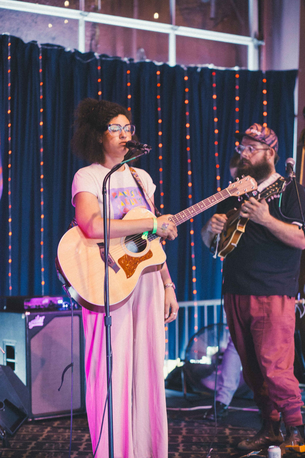 Two musicians perform on stage, one singing and playing guitar, the other playing guitar, with a dark curtain backdrop and stage lights.