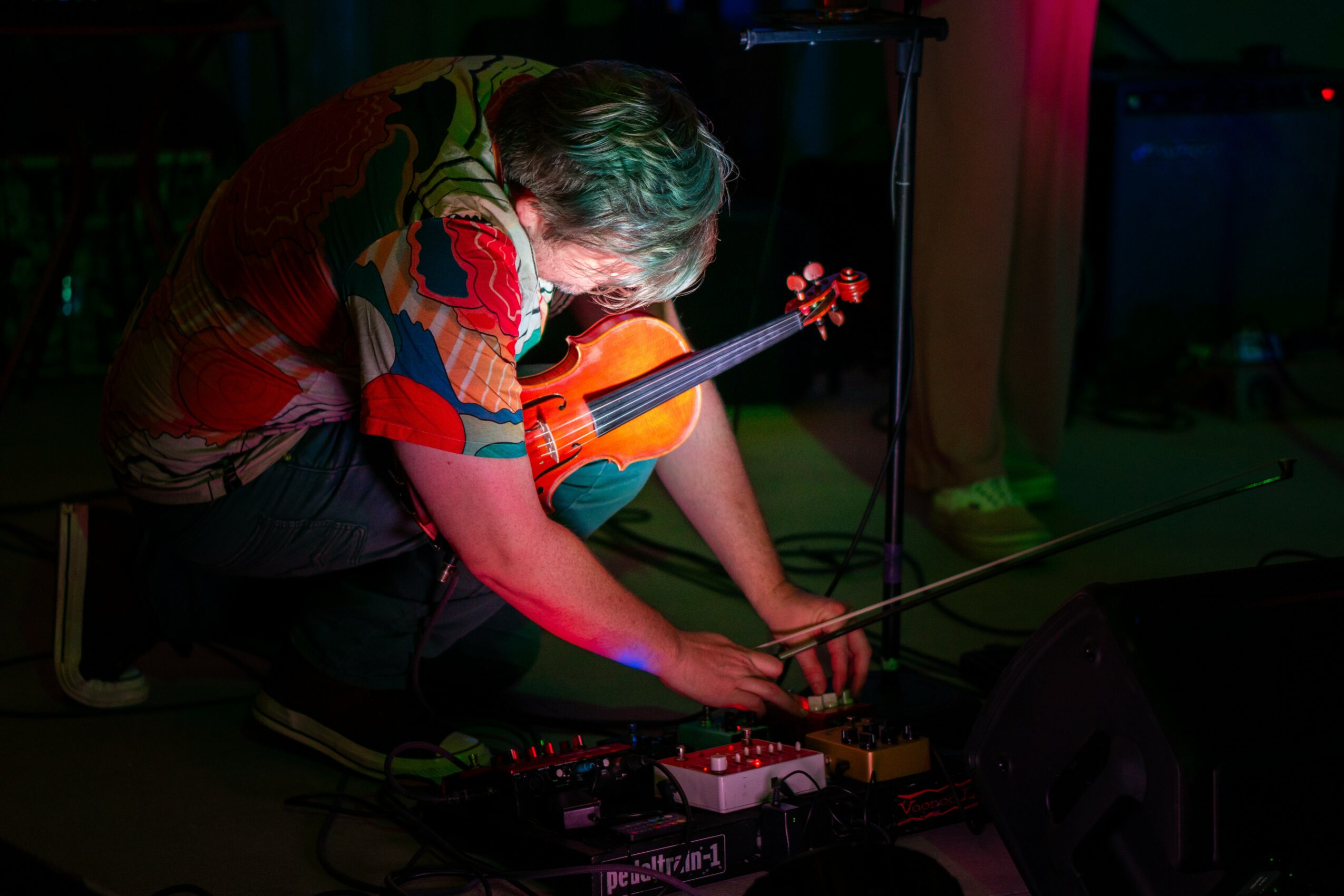 Person crouching on the floor adjusting musical equipment with a guitar nearby, in a dimly lit room.