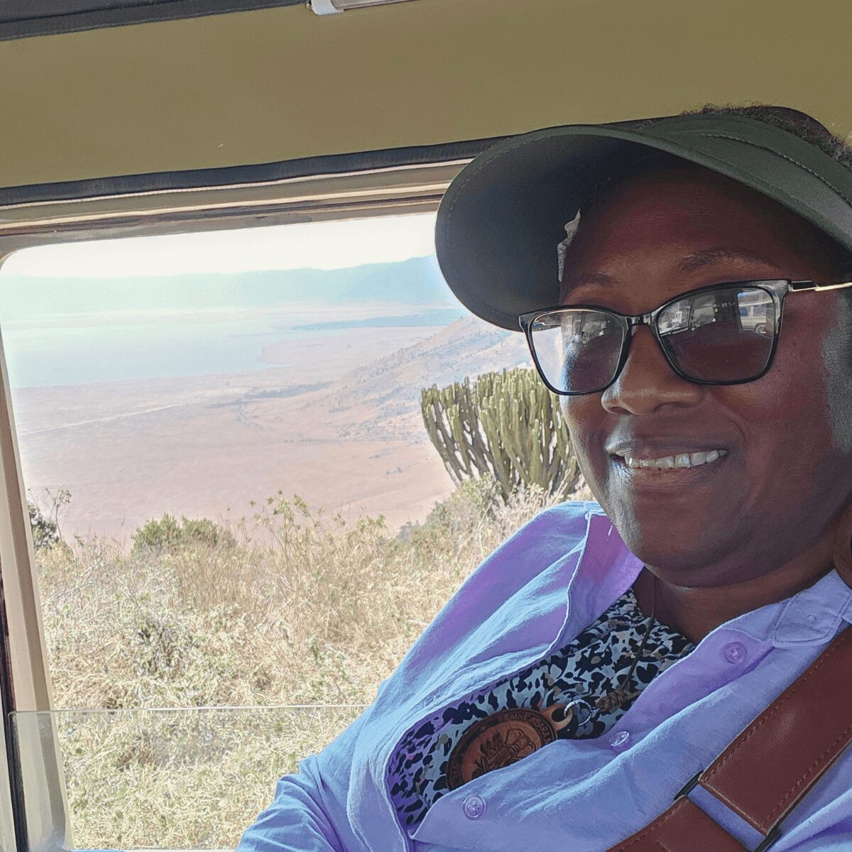 Person wearing glasses and a hat, smiling, with a desert landscape and cactus outside window.