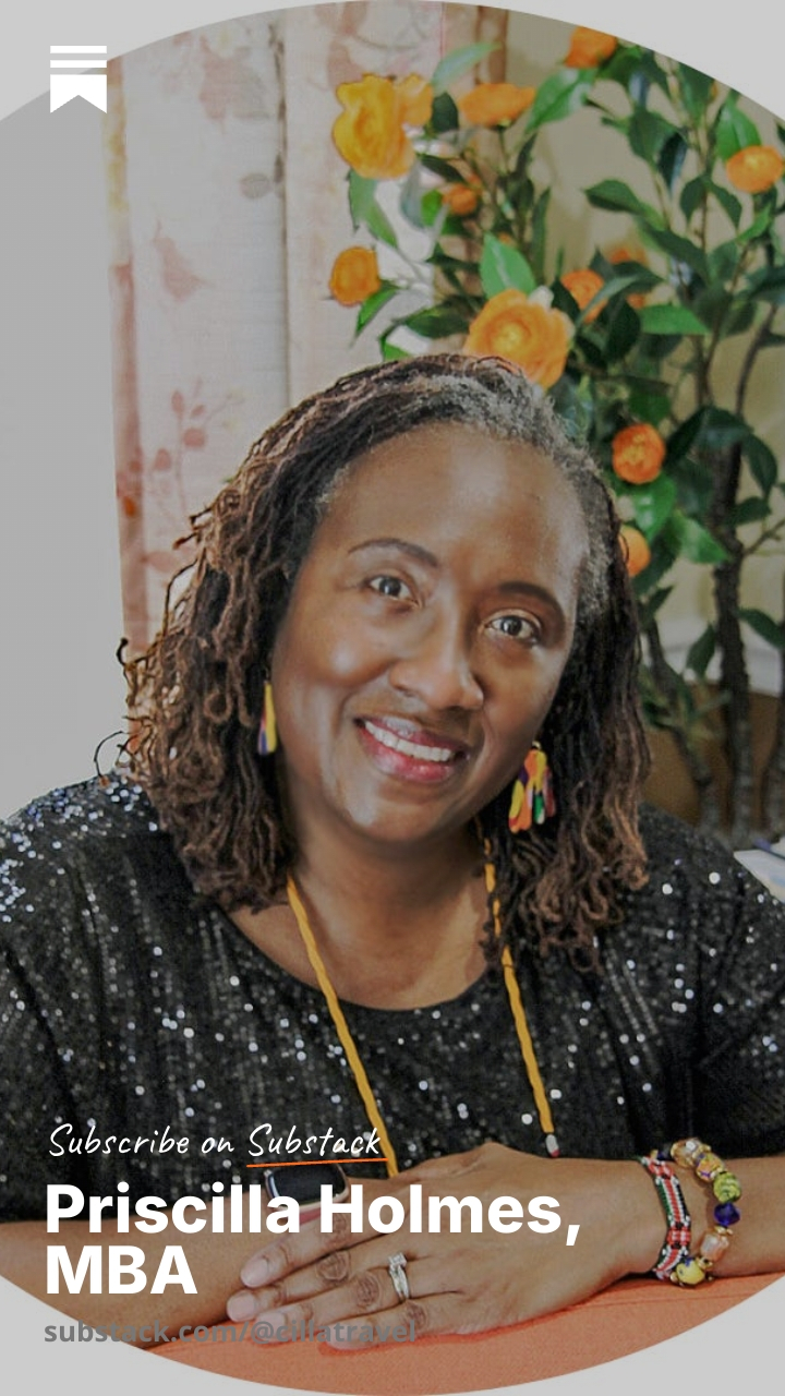 Smiling woman with curly hair wearing a black sequined top, jewelry, and sitting at a table with flowers in background.