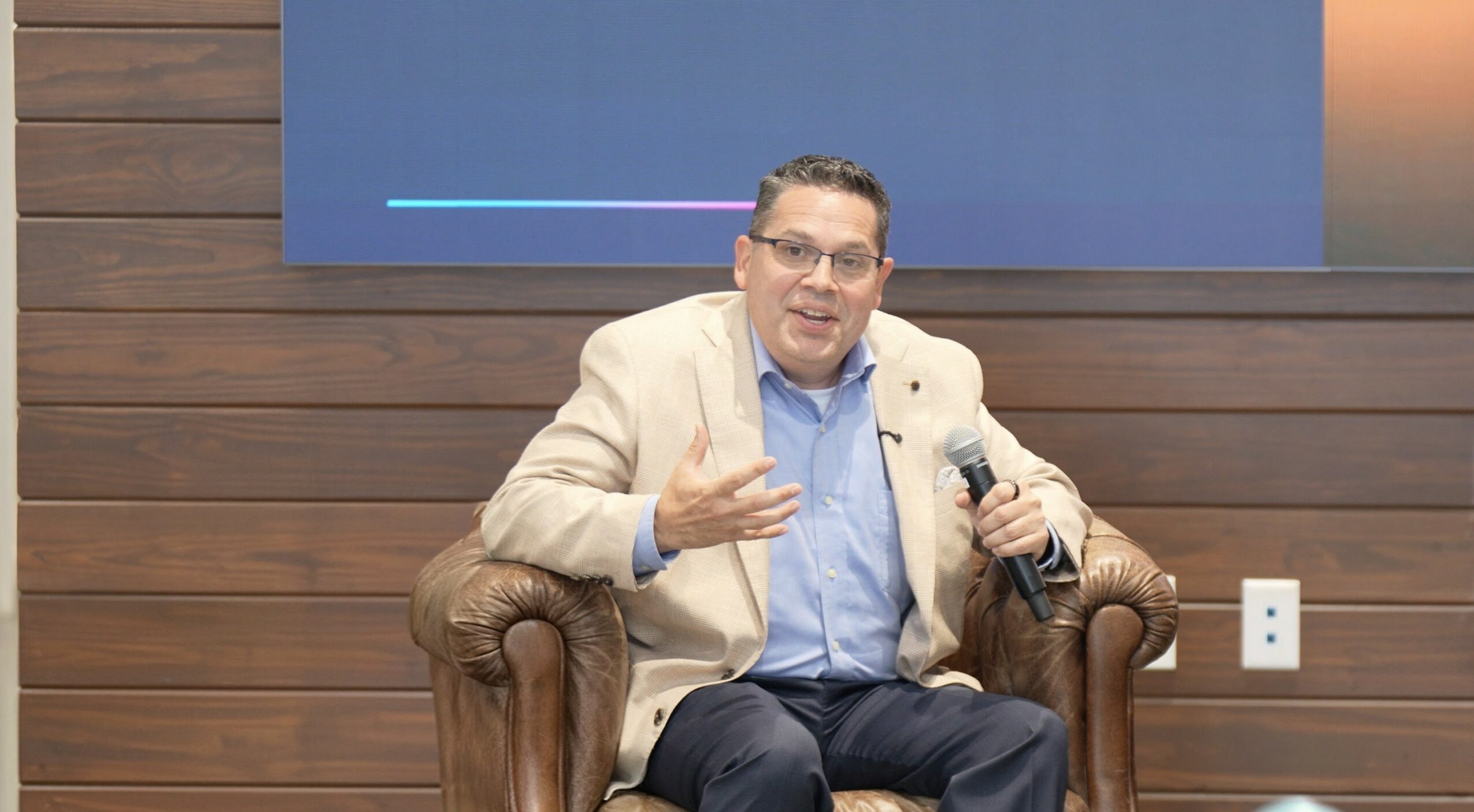 Man in beige blazer and blue shirt sitting on a leather armchair, speaking and gesturing with his hand, in front of a wooden wall.