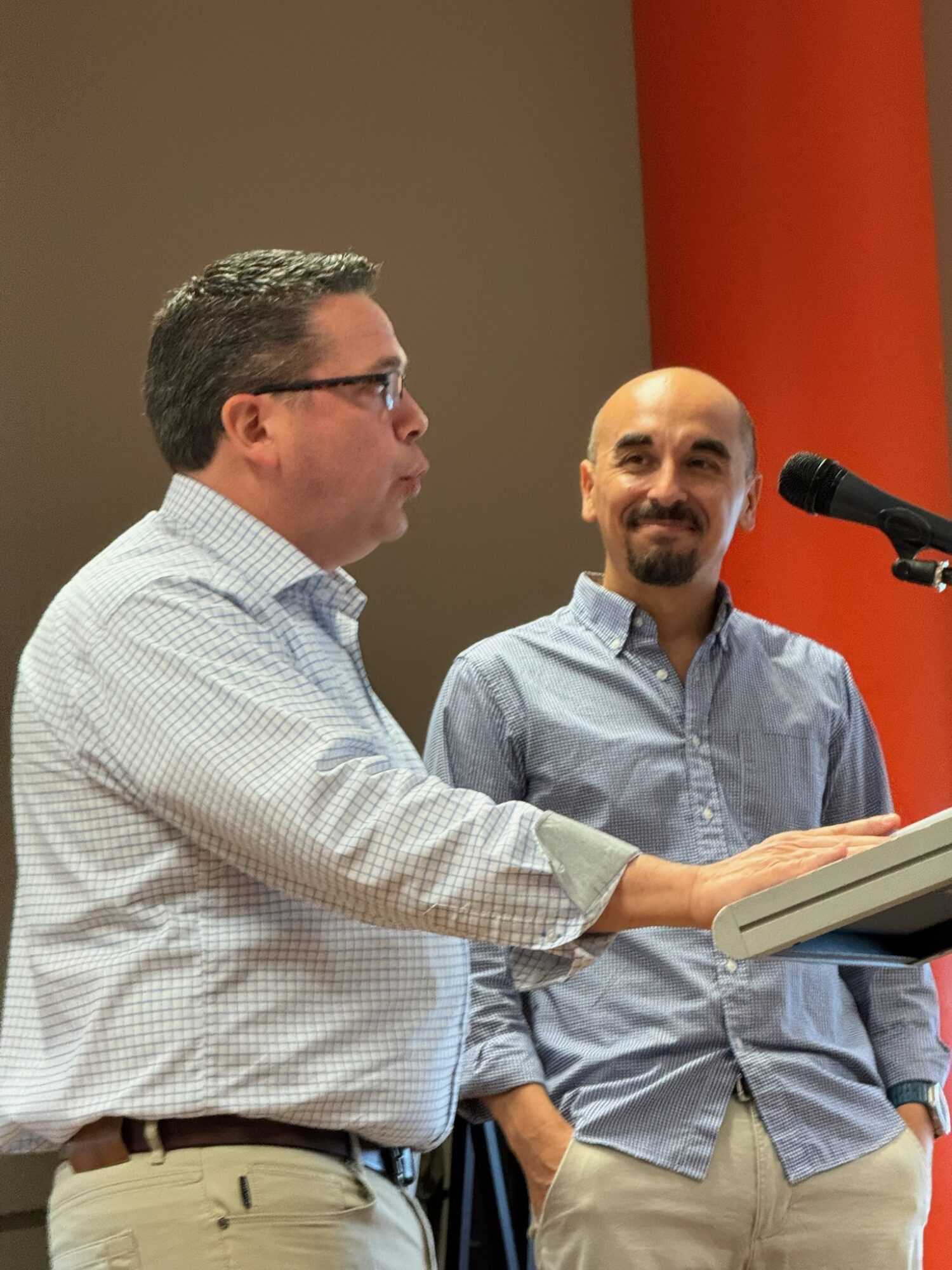 Two men standing at a podium, one speaking and the other smiling, in front of a red and beige background.