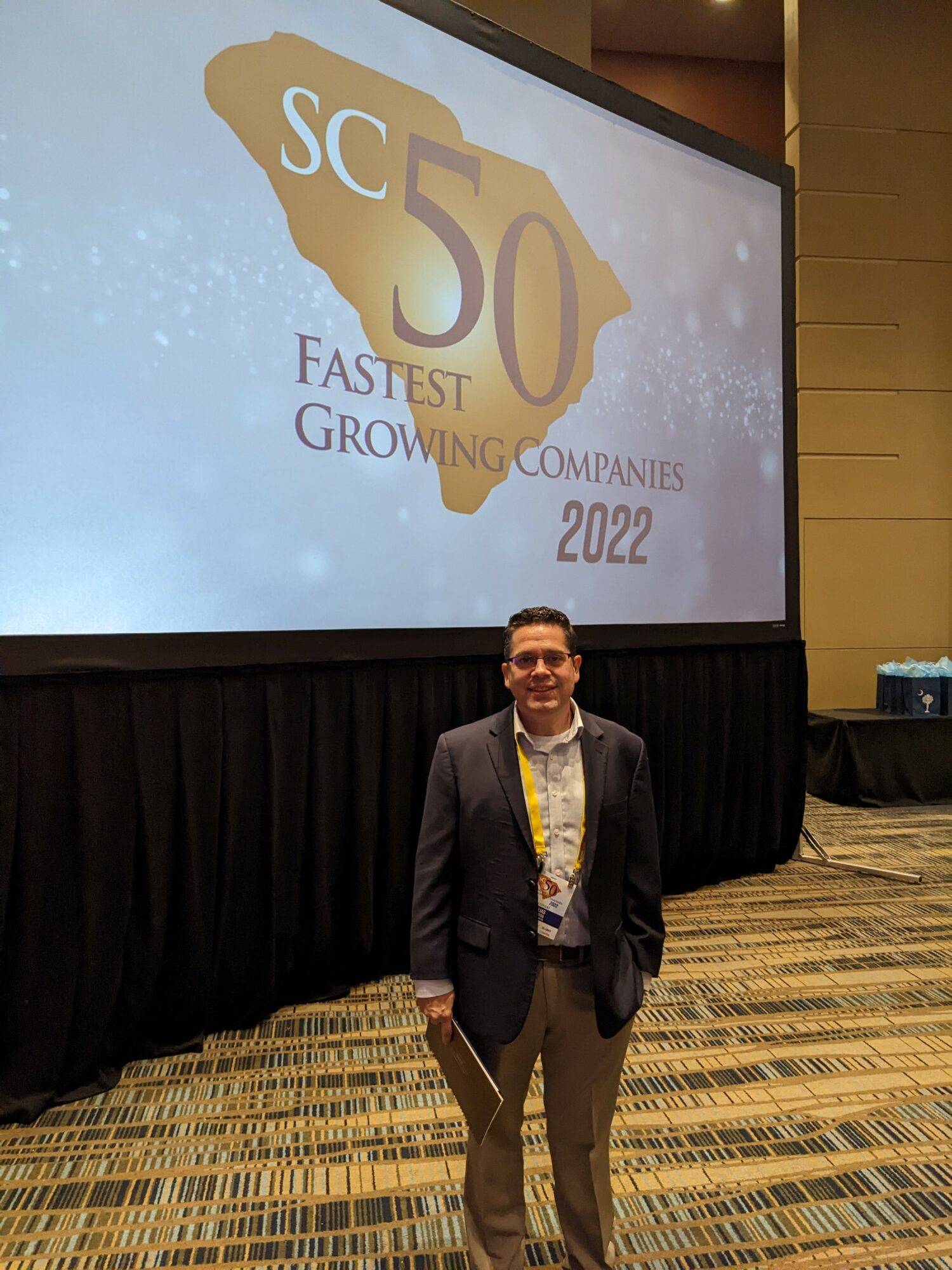 Man in suit standing in front of a large screen displaying a logo with the text 'SC 50 Fastest Growing Companies 2022' in a conference room.