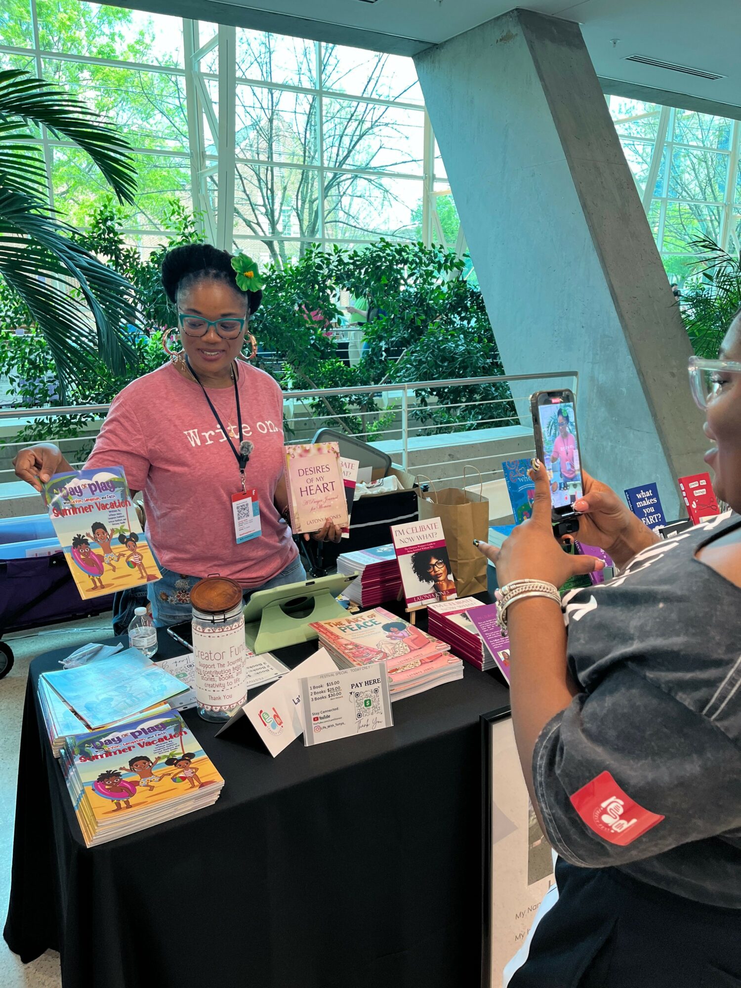 Person standing at a table with books and promotional materials, with a glass window and greenery outside in the background.