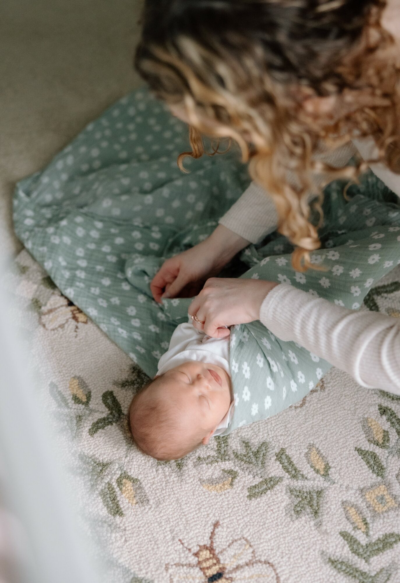 Person gently holding a newborn baby on a patterned rug, baby wrapped in a polka dot blanket, person wearing a light-colored long sleeve.
