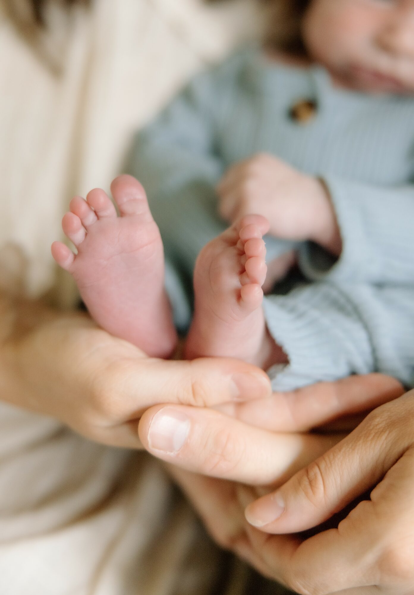 Close-up of a person's hand holding a baby's feet, with the baby wearing a light-colored outfit, blurred background.