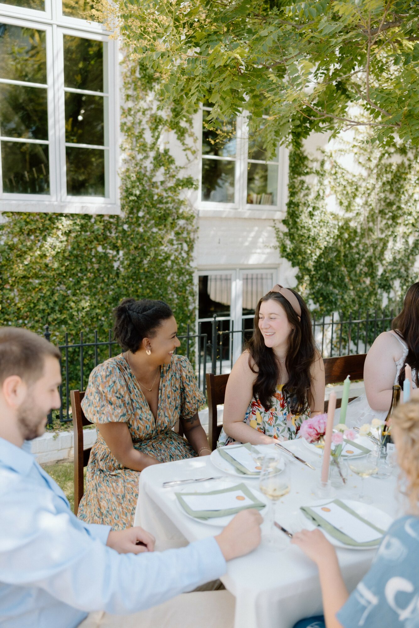 People sitting at a table outdoors, smiling and talking, with a building and greenery in the background.