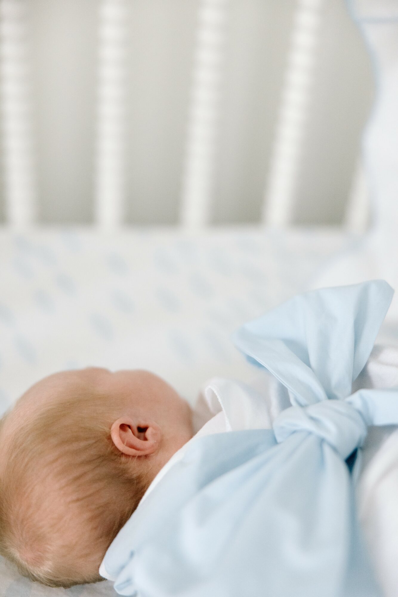 Close-up of a sleeping baby lying in a crib, wrapped in a light blue blanket, with a white crib railing in background.