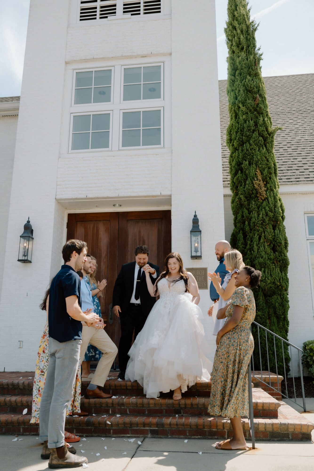 Group of people, including a bride in a wedding dress, standing on steps outside a white building with tall tree.