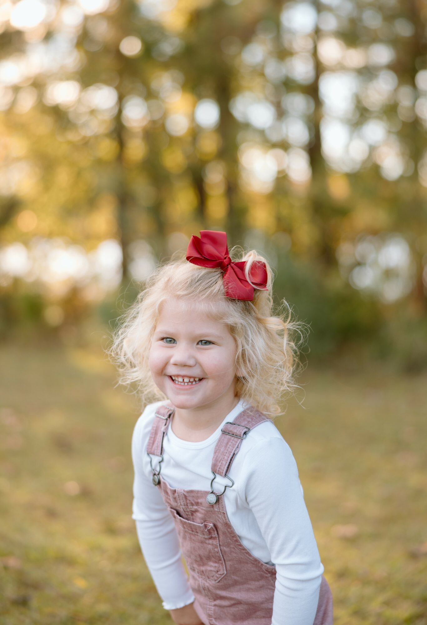 Young girl with curly blonde hair, red bow, smiling outdoors with trees in background.