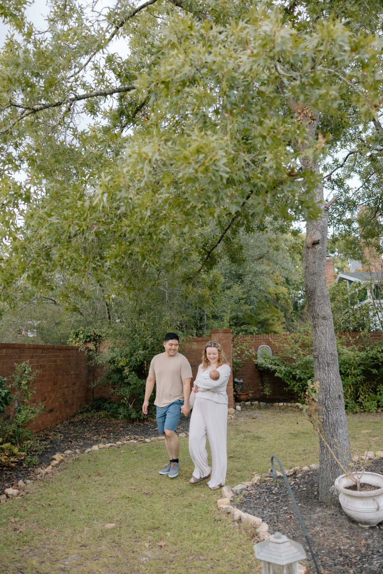 Two people walking in a garden with trees and a brick wall, one holding a phone, sunny day.
