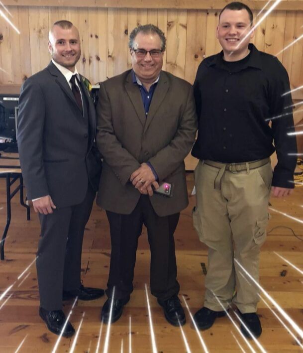 Three men standing indoors in front of a wooden wall, smiling, dressed in formal and casual attire.
