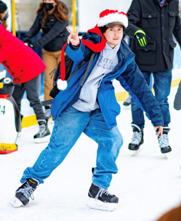 Child wearing a Santa hat and blue jacket ice skating, making a peace sign with one hand, surrounded by other skaters.