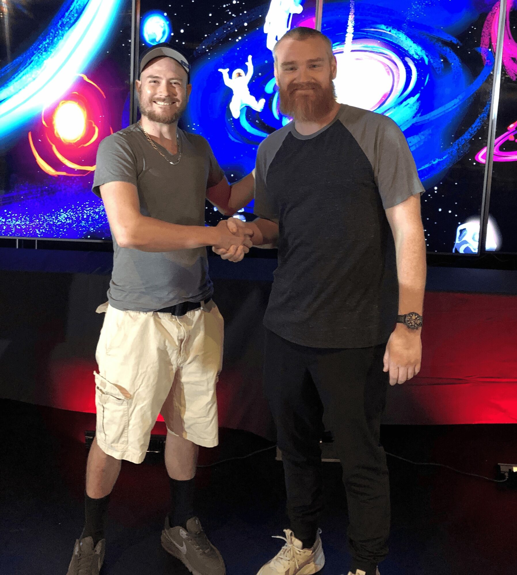 Two men shake hands in front of a colorful space-themed digital background, smiling at the camera.