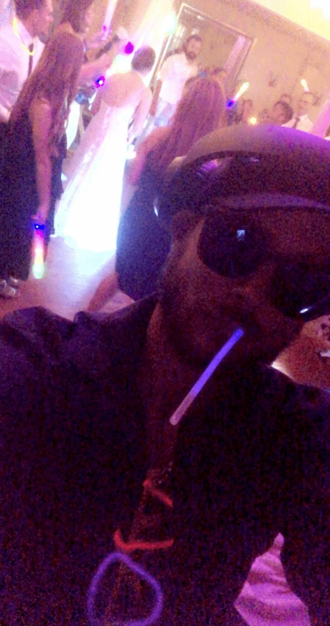 Person wearing sunglasses and a cap taking a selfie at a party with people dancing in the background.
