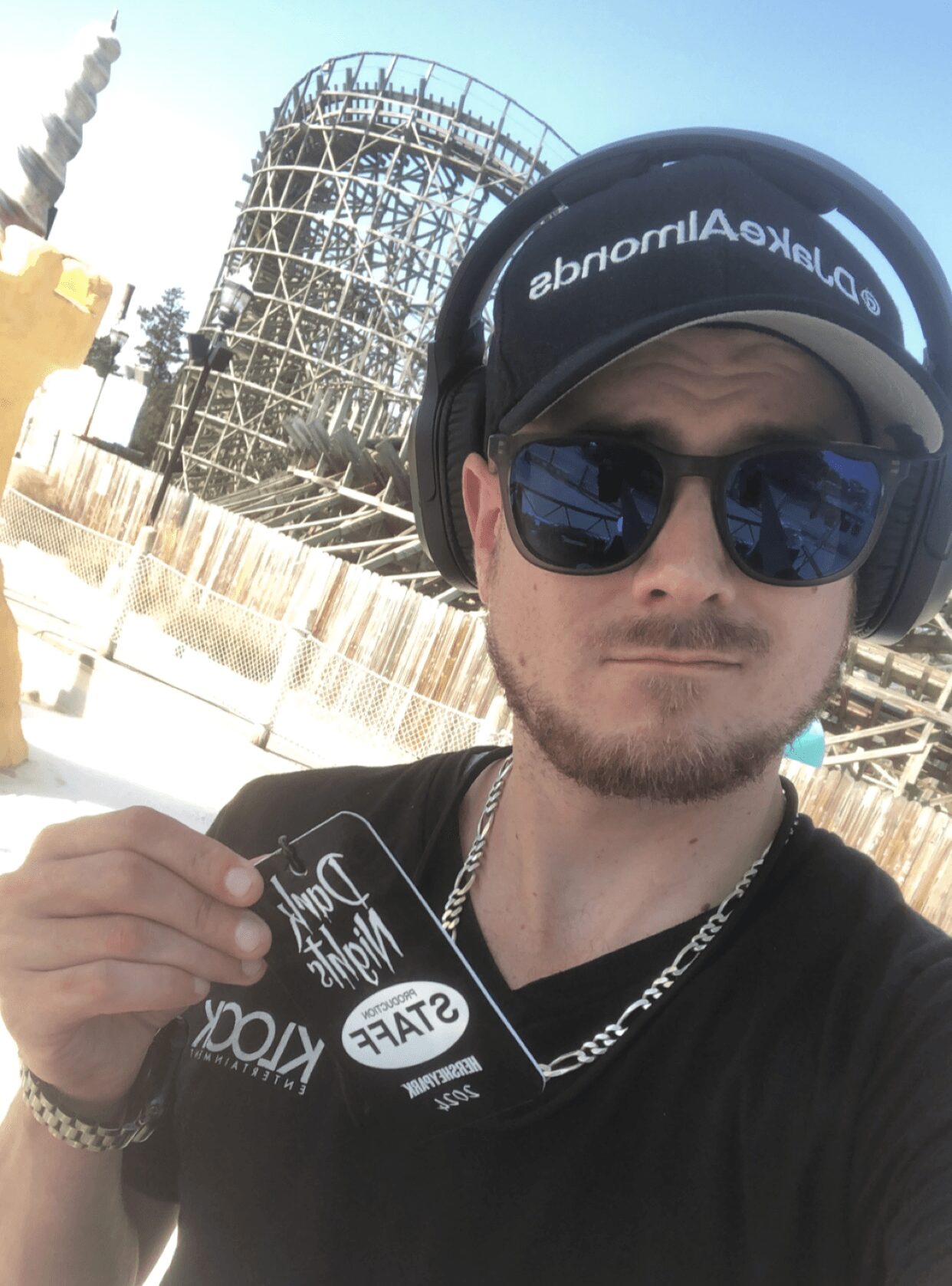 Young man wearing headphones and sunglasses taking a selfie outdoors with a roller coaster in the background.