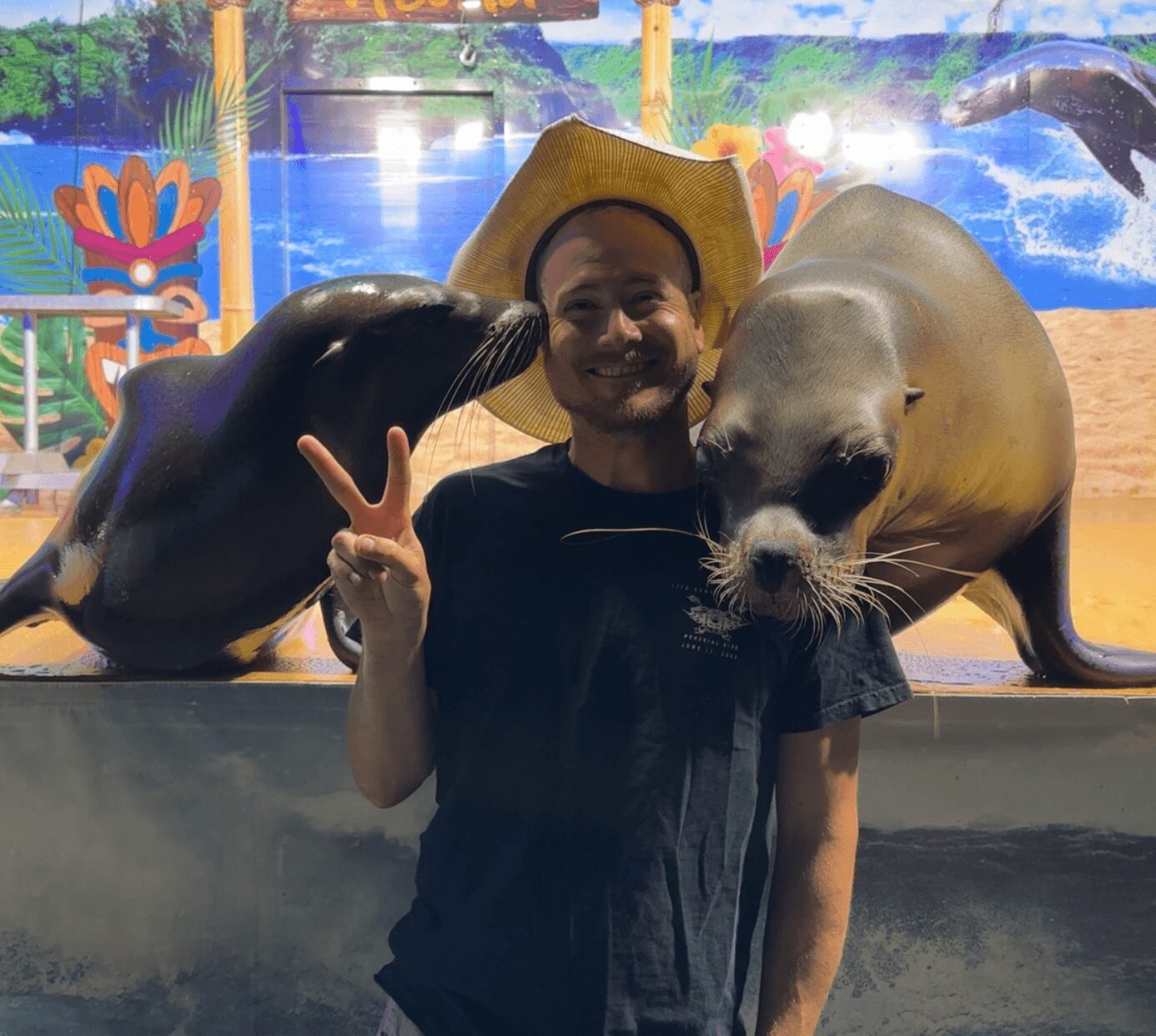 Person wearing a cowboy hat making a peace sign with two sea lion heads close to their face, colorful background.