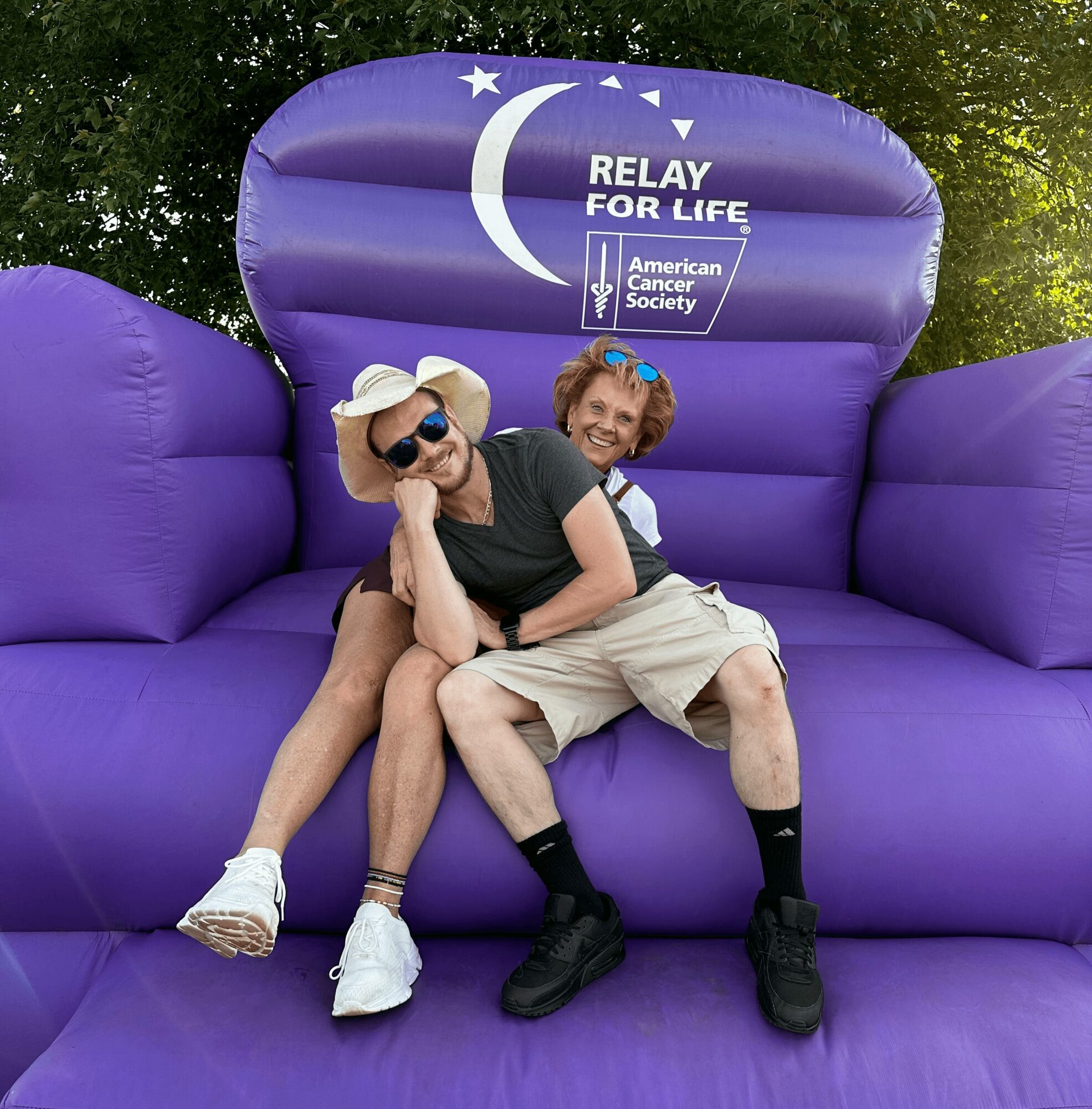 Two smiling people sitting on a large purple inflatable chair with a sign for Relay for Life and American Cancer Society, outdoors.