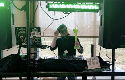 Person wearing sunglasses and headphones, holding a green object, standing behind DJ equipment with speakers and a window in background.
