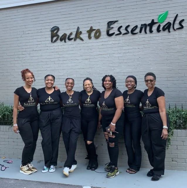 Group of six women standing in front of a wall with 'Back to Essentials' sign, smiling, wearing matching black shirts, and casual pants.