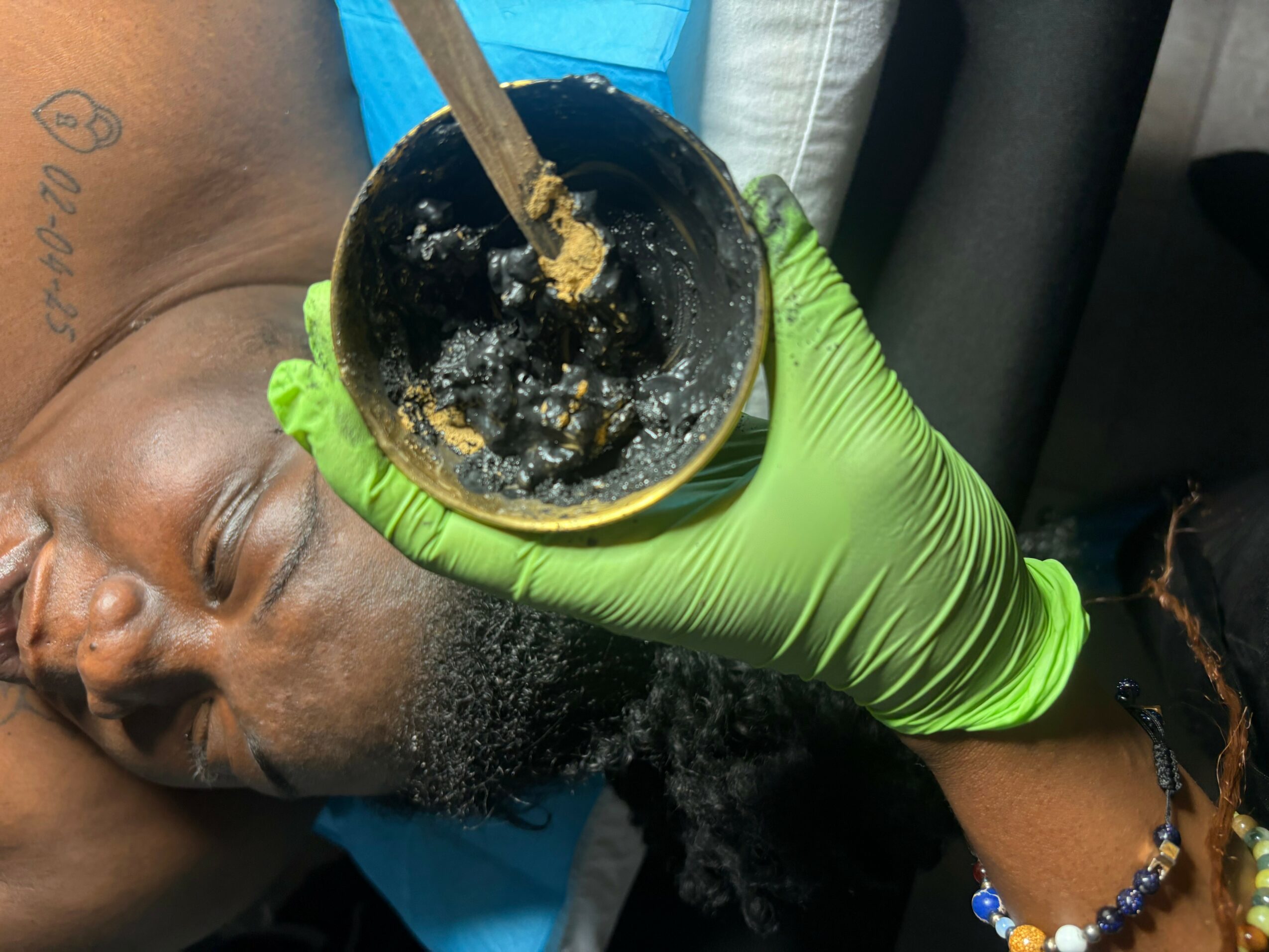 Person lying down with eyes closed, holding a container with a spoon, wearing a green glove, in a vehicle.