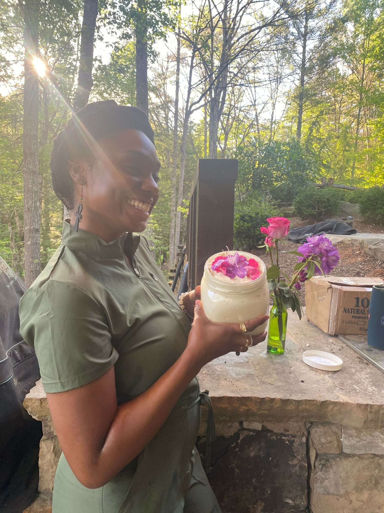 Person holding a drink with pink flowers, outdoors with trees and flowers in background.