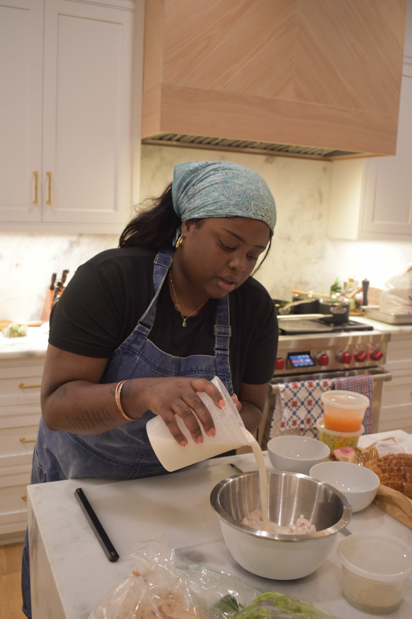 Woman wearing a headscarf and apron preparing food in a kitchen, with various ingredients and utensils on the table.
