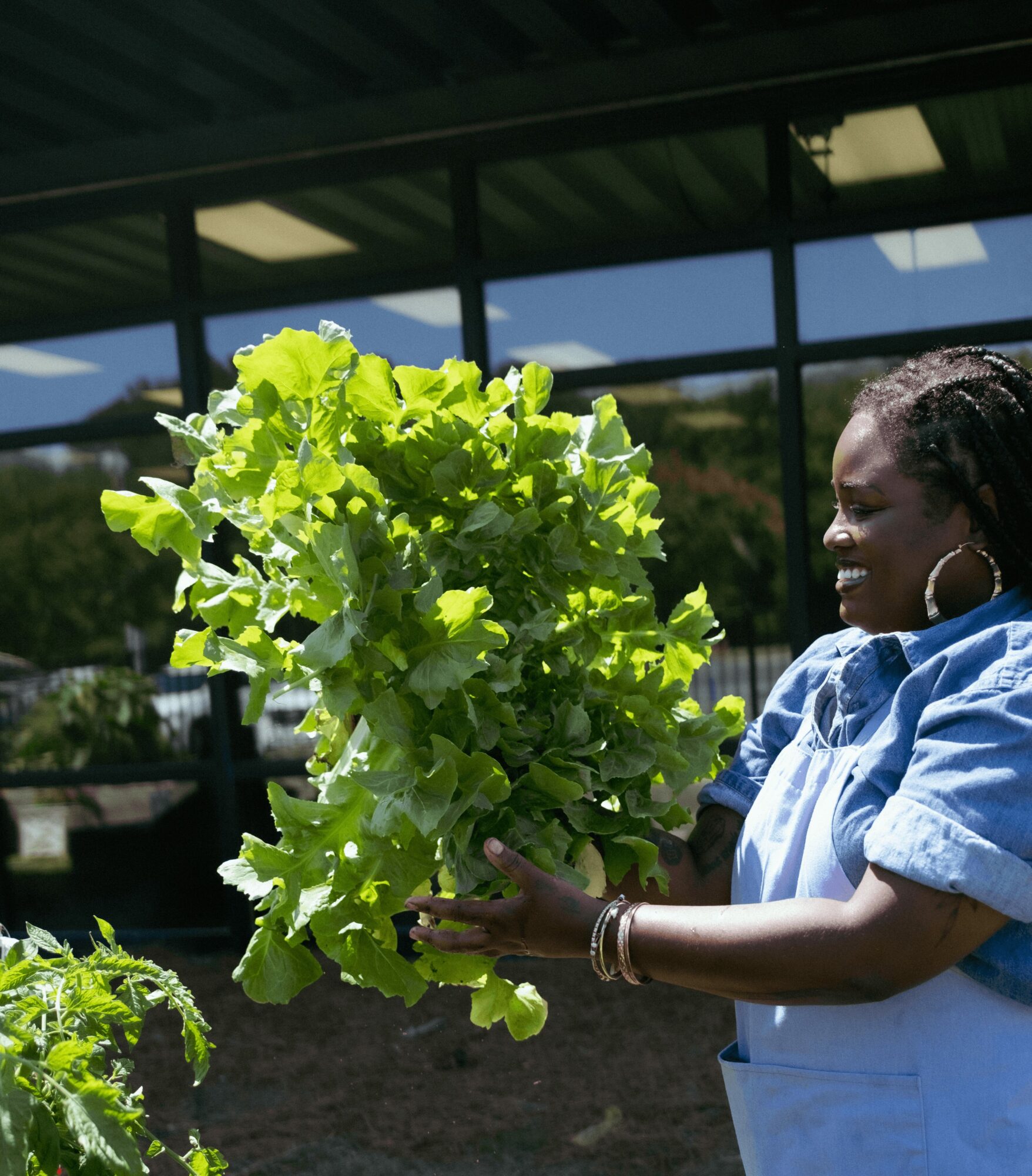 Woman holding a large bunch of green lettuce outdoors, smiling, with a building and trees in the background.