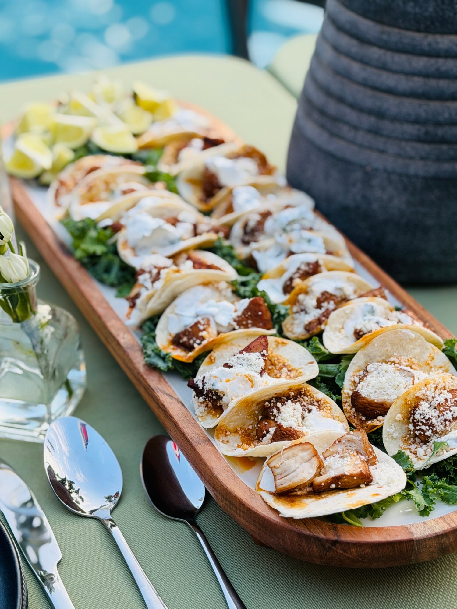 A long wooden platter with assorted tacos, garnished with lime wedges and greens, on a table with spoons nearby.
