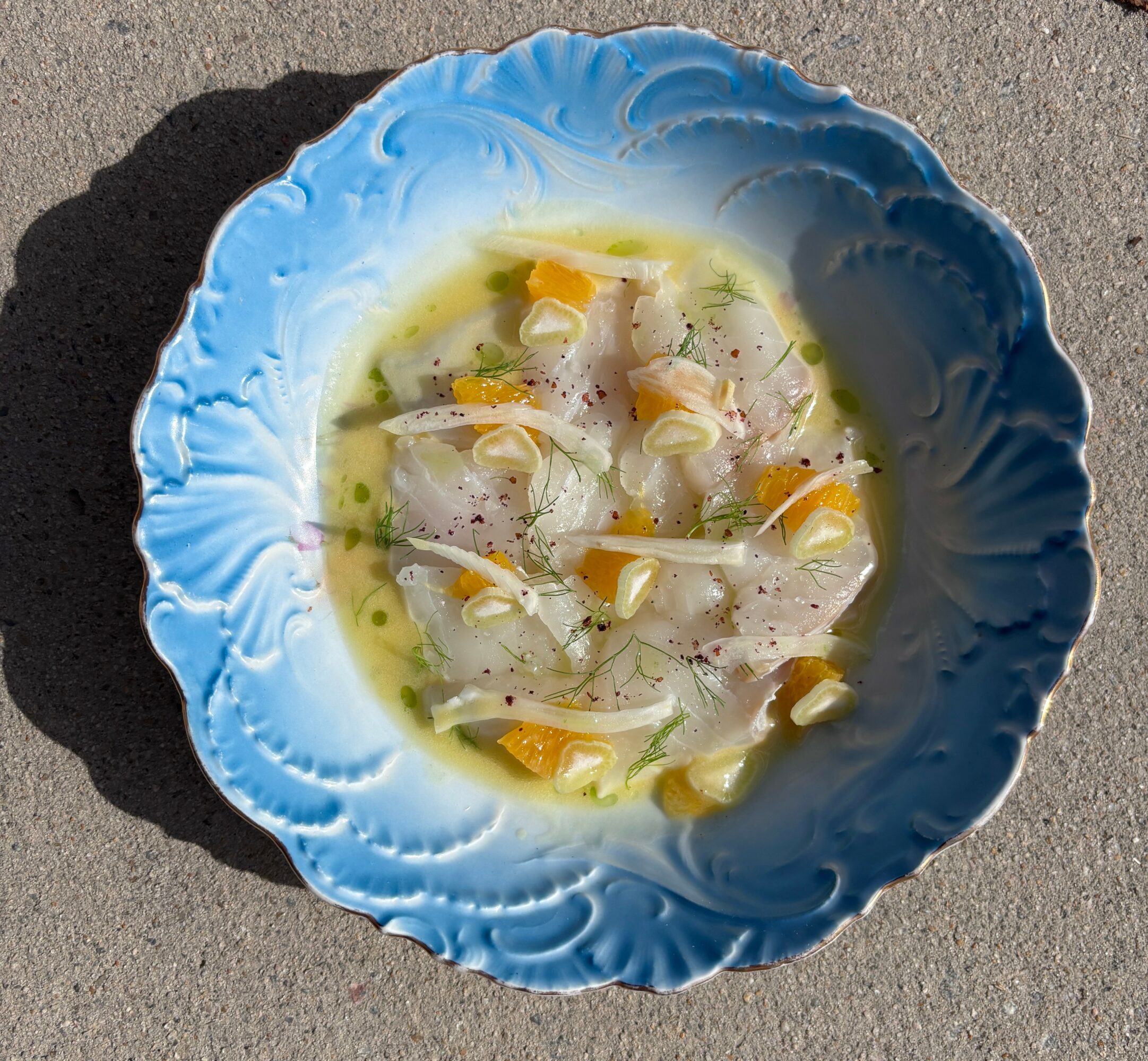 Bowl of fish ceviche with corn, onions, and herbs on a gray surface, sunlight casting a shadow.
