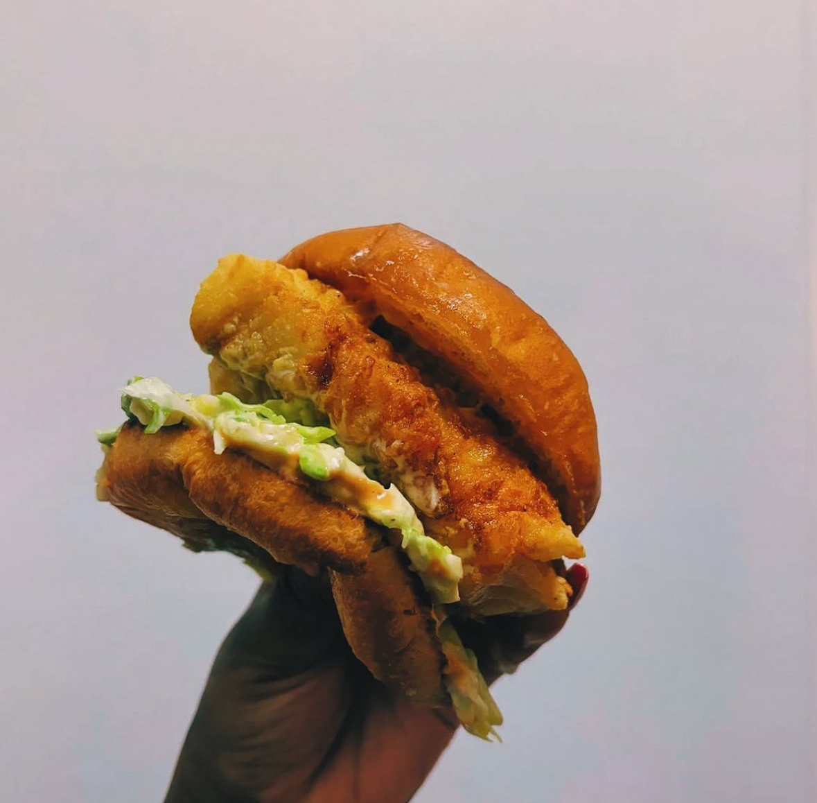 Hand holding a fried chicken sandwich with lettuce and sauce, against a plain background.