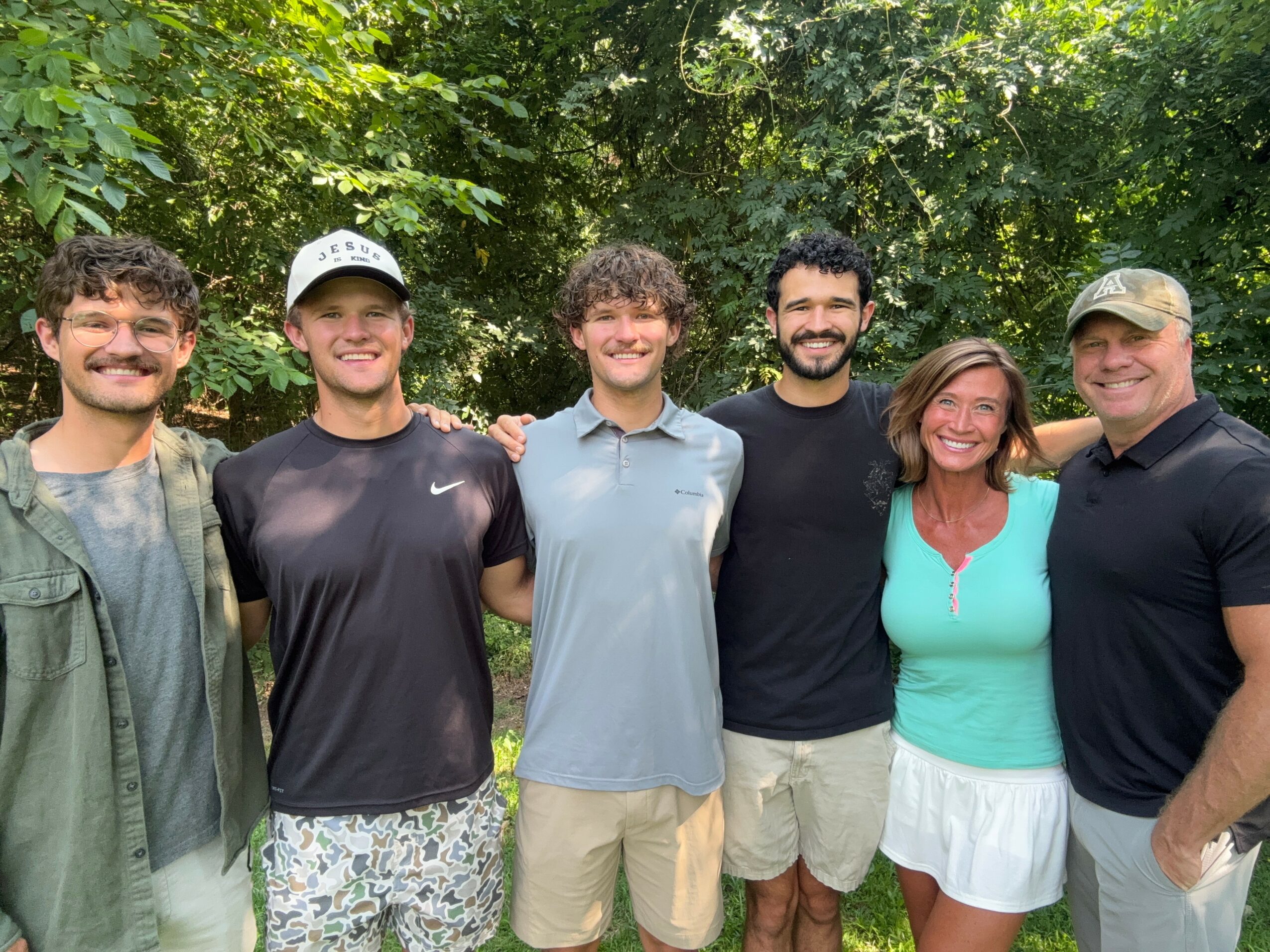 Six people standing outdoors in front of trees, smiling, with arms around each other, dressed casually.