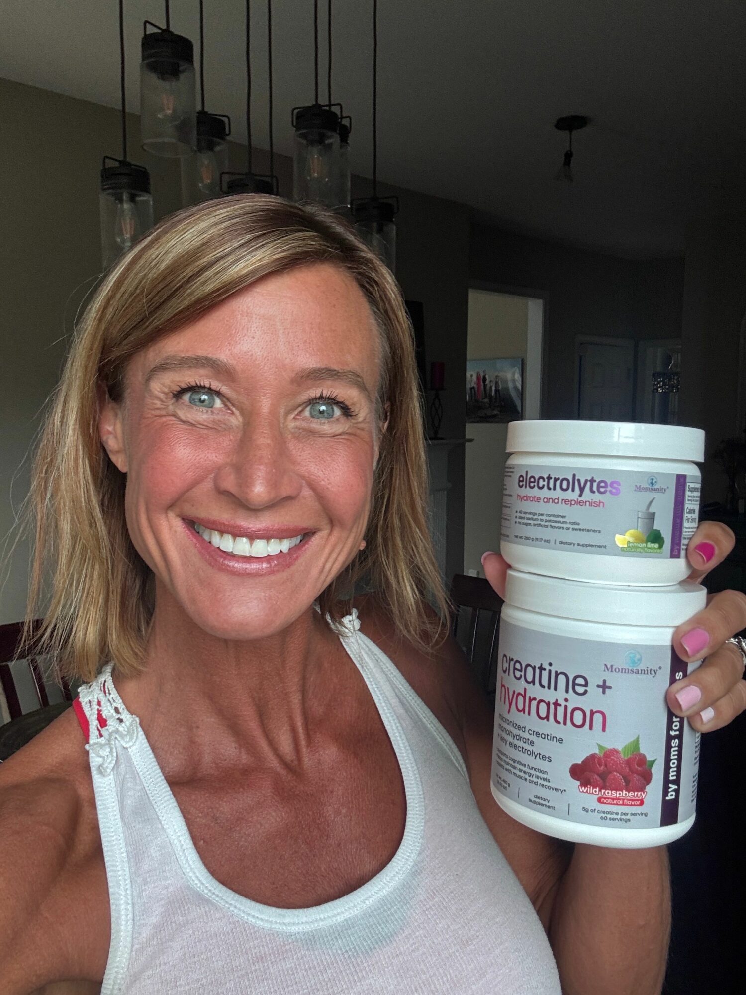 Woman with blonde hair smiling, holding a bottle of dietary supplement, wearing a white tank top.