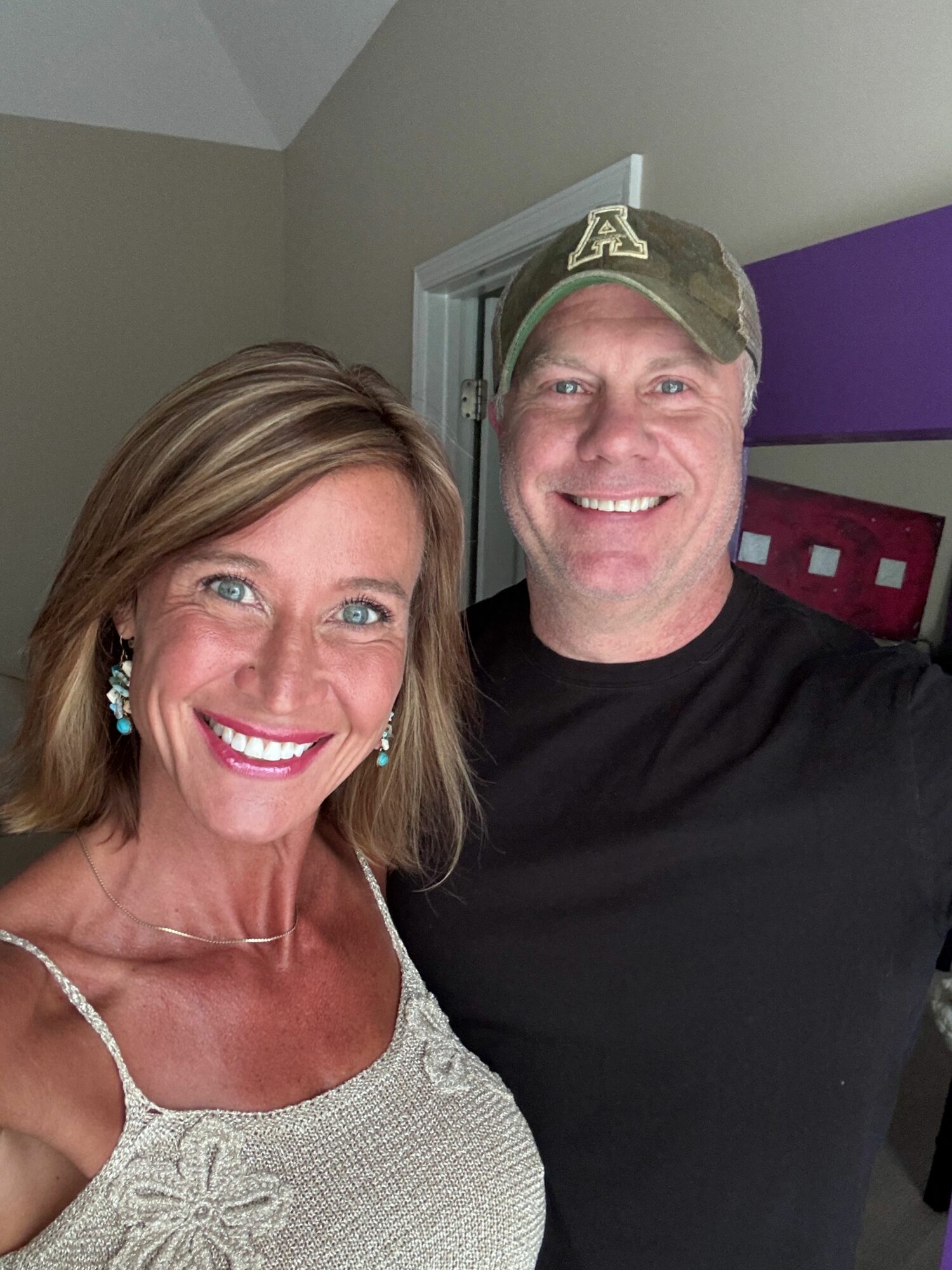 Two smiling people, a man and a woman, pose indoors with a purple wall in the background.